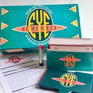 May include: The Eye Remember board game set includes a large teal box with the title text, a smaller teal card holder, a stack of game cards, a clear plastic hourglass with a red cap, and a white instruction sheet with black printed text.