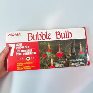 以下が含まれることがあります： 長方形の箱に入ったNOMAバブルバルブ7灯屋内用ライトセット。パッケージは白と赤で、Bubble Bulb、7 light indoor set、bubbles continuously when litなどの文字が書かれています。箱には赤と緑のクリスマスライトの画像が印刷されています。