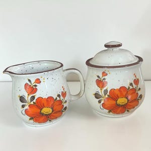 Peut inclure: Un pot à lait et un sucrier avec couvercle en céramique, présentant un émail blanc cassé moucheté orné de fleurs orange peintes à la main, de cœurs jaunes et de tiges brunes. Les deux pièces ont un bord brun et une forme arrondie.