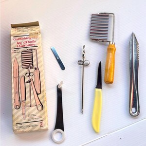 Peut inclure: Un ensemble de cinq ustensiles de cuisine pour garniture comprenant un coupe-frites ondulé avec manche en bois, un petit couteau d'office à manche jaune, un pic à garniture, un outil à boucle et une lame plate, présentés dans une boîte en carton.