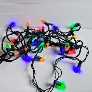 以下が含まれることがあります： 赤、緑、オレンジ、青、黄色のファセット加工された電球型カバーが付いたマルチカラーのLEDクリスマスライトのストリング。ライトはダークグリーンのワイヤーで接続されており、光る電球が絡み合った状態になっています。