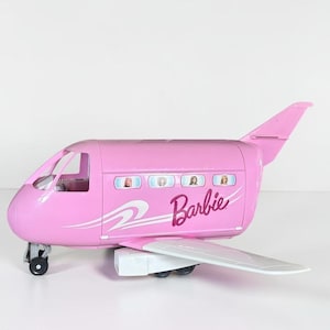 Puede incluir: Un avión de juguete rosa con alas y tren de aterrizaje blancos. El lateral del fuselaje presenta la palabra Barbie en letra cursiva, líneas gráficas blancas y cuatro pequeñas ventanas que muestran imágenes de muñecas en la cabina.