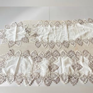 Peut inclure: Un ensemble de chemins de table en tissu blanc cassé et un napperon carré assorti, chacun doté de bordures en dentelle beige festonnée complexe avec des motifs floraux et géométriques détaillés sur les bords, formant une collection de linge de table coordonnée.