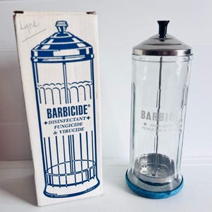 Puede incluir: Un frasco desinfectante Barbicide de vidrio transparente con tapa de acero inoxidable, pomo negro y base azul, acompañado de su caja de cartón blanca original con gráficos azules y el texto Barbicide Disinfectant Fungicide & Virucide.