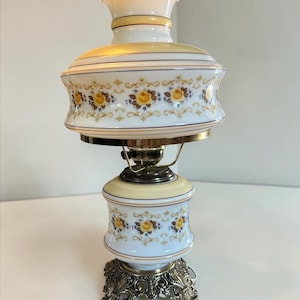 Peut inclure: Une lampe de table avec un abat-jour et une base en verre opalin blanc, ornés de motifs de roses jaunes et de détails en filigrane doré. La lampe présente un bord supérieur festonné, une base en métal doré et une monture centrale en laiton.