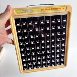 May include: A vintage Touch 'n Tell Me educational toy with a yellow plastic frame and a black grid panel featuring white buttons for addition and subtraction math problems. The top label displays the text Touch 'n Tell Me and Addition & Subtraction.
