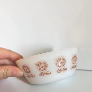 Peut inclure: Un bol en verre opalin blanc orné d'un motif répétitif de fleurs carrées marron stylisées avec des tiges et des feuilles. Le bol présente un bord arrondi lisse et une base légèrement effilée, offrant une finition opaque et uniforme.