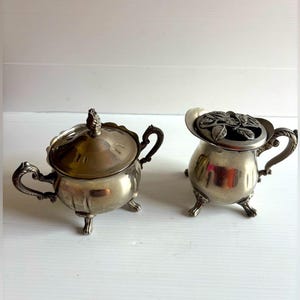 Peut inclure: Deux pièces de service antiques en argent plaqué. L'une est un sucrier couvert avec un bouton décoratif et des poignées ornées. L'autre est une crémière avec un motif floral sur le couvercle et des poignées assorties. Les deux pièces ont de petits pieds.