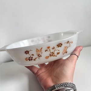 Peut inclure: Un plat de cuisson ovale en verre blanc orné d'un motif répétitif de papillons et de branches fleuries marron. Le plat est doté de poignées intégrées aux deux extrémités et présente une finition lisse et brillante.