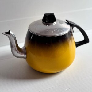 Puede incluir: Una tetera vintage con un diseño degradado de amarillo a negro. Cuenta con una tapa y un pico plateados, y un asa negra. La tetera está diseñada para preparar té.