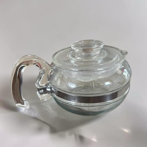 Peut inclure: Une théière en verre transparent dotée d'un corps arrondi, d'un couvercle en verre assorti avec une poignée en bouton et d'une anse latérale en verre incurvée. Une bande métallique argentée entoure le milieu de la théière pour la fixation.