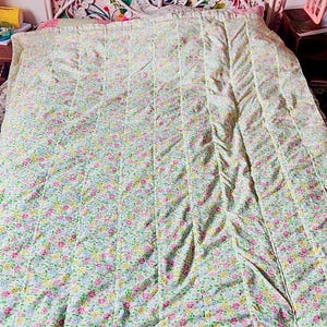Peut inclure: Un couvre-lit matelassé présentant un motif floral répétitif avec de petites fleurs roses, jaunes et bleues sur un fond blanc. Le tissu est cousu avec des canaux verticaux sur toute la longueur, créant un aspect texturé et linéaire.
