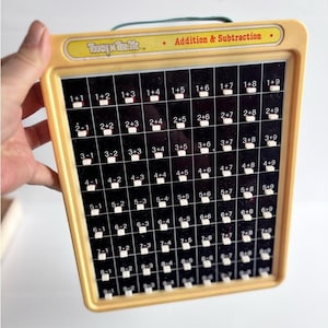May include: A vintage Touch 'n Tell Me educational toy with a yellow plastic frame and a black grid panel featuring white buttons for addition and subtraction math problems. The top label displays the text Touch 'n Tell Me and Addition & Subtraction.
