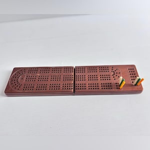 Peut inclure: Un plateau de cribbage en plastique marron doté d'un design pliable avec une grille de trous pour le score. Le plateau comprend plusieurs petits piquets fins de couleurs blanche, verte et orange insérés dans les trous sur le côté droit.