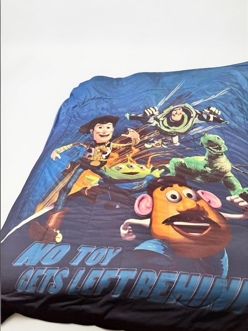 Puede incluir: Una colcha azul con personajes de la pel&iacute;cula de animaci&oacute;n Toy Story. El dise&ntilde;o incluye a Woody, Buzz Lightyear, Rex, los Aliens y el Sr. Potato. El texto "NO TOY GETS LEFT BEHIND" est&aacute; impreso en la parte inferior.