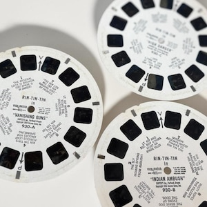 May include: Three vintage View-Master reels with black and white images and text. The reels feature the text "Rin-Tin-Tin" and titles like "Vanishing Guns" and "Indian Ambush". Each reel has a series of small, square viewing windows.