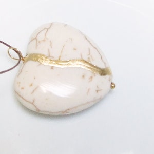 May include: A white heart-shaped pendant with gold accents. The pendant has a natural, marbled look and is strung on a brown cord.