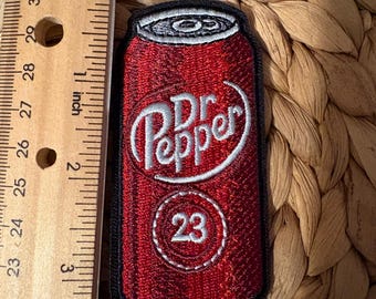 Op de afbeelding: Een geborduurde patch in de vorm van een rood Dr Pepper-blikje met witte tekst en het witte getal 23 in een cirkel. De patch ligt naast een houten liniaal die een totale hoogte van ongeveer 8,9 cm aangeeft.