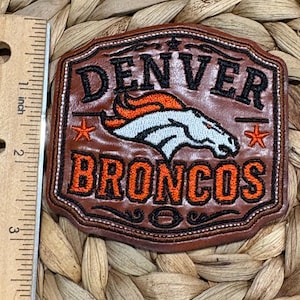 May include: A brown leather patch featuring the Denver Broncos logo, the text "DENVER" and "BRONCOS" in black and orange embroidery, and two orange stars. A ruler placed to the left shows the patch is approximately 2.5 inches in height.