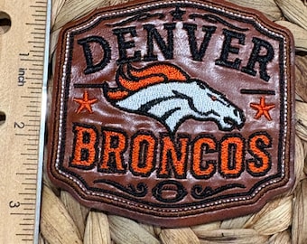 Op de afbeelding: Een bruine leren patch met het Denver Broncos logo, de tekst "DENVER" en "BRONCOS" in zwart en oranje borduursel, en twee oranje sterren. Een liniaal aan de linkerkant toont dat de patch ongeveer 6,4 cm hoog is.
