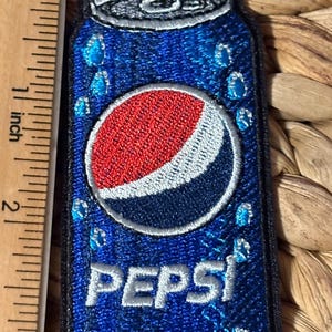 May include: A rectangular embroidered patch shaped like a blue soda can with the Pepsi logo in red, white, and blue. The patch features light blue water droplets and the text PEPSI in white. A wooden ruler shows the patch is 3 inches long.