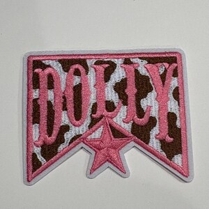 May include: A rectangular embroidered patch with a brown and white cow print background. The word DOLLY is stitched in pink thread across the center, positioned above a pink five-pointed star. The patch has a white border and a pointed bottom edge.