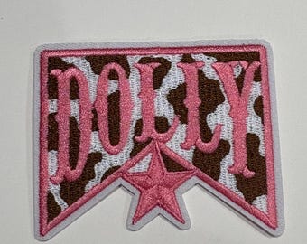 Op de afbeelding: Een rechthoekige geborduurde patch met een bruin-witte koeienprint als achtergrond. Het woord DOLLY is in roze draad in het midden geborduurd, boven een roze vijfpuntige ster. De patch heeft een witte rand en een puntige onderkant.