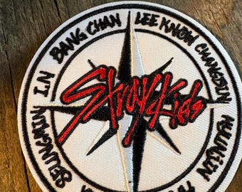 Op de afbeelding: Een ronde geborduurde patch met een witte achtergrond en een zwart kompassterontwerp. De rode tekst Stray Kids staat gecentreerd over de ster. De namen Bang Chan, Lee Know, Changbin, Hyunjin, Han, Felix, Seungmin en I.N zijn in het zwart langs de buitenrand geborduurd.