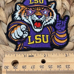 Op de afbeelding: Een geborduurde patch met een tijgermaskotte die een paarse pet met de tekst LSU, stervormige brillen en een paars shirt met de tekst LSU draagt. De tijger wijst naar voren en maakt een handgebaar. Een houten liniaal toont aan dat de patch ongeveer 7,6 cm breed is.