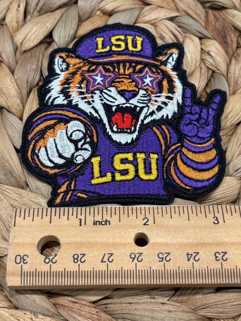 Op de afbeelding: Een geborduurde patch met een tijgermaskotte die een paarse pet met de tekst LSU, stervormige brillen en een paars shirt met de tekst LSU draagt. De tijger wijst naar voren en maakt een handgebaar. Een houten liniaal toont aan dat de patch ongeveer 7,6 cm breed is.