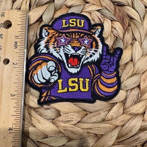May include: An embroidered patch featuring a tiger mascot wearing a purple cap with the text LSU, star-shaped glasses, and a purple jersey with the text LSU. The patch is next to a ruler showing a height of approximately 3.5 inches.