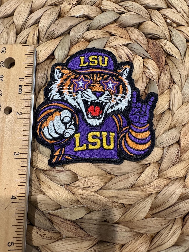 Op de afbeelding: Een geborduurde patch met een tijgermaskotte die een paarse pet met de tekst LSU, stervormige brillen en een paars shirt met de tekst LSU draagt. De patch ligt naast een liniaal die een hoogte van 8,9 cm aangeeft.