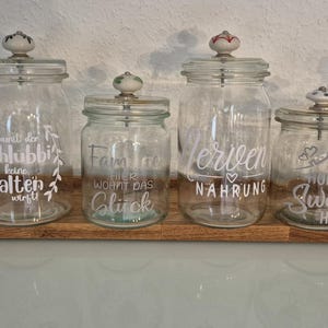 Könnte beinhalten: Sechs durchsichtige Glasbehälter mit dekorativen Deckeln und weißer Beschriftung. Die Behälter sind mit folgendem Text beschriftet: "Geheim Vorrat", "Daunlt ein Schlubbie", "Familie wohnt das Glück", "Verlieben Nahrung", "Home Sweet Home" und "Süßigkeiten ern do in sp". Die Behälter stehen auf einer Holzoberfläche.