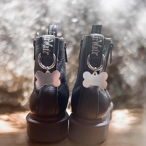 Könnte beinhalten: Schwarze Leder-Stiefeletten mit einem silbernen Hundefell-Anhänger an jedem Stiefel. Die Stiefel haben einen seitlichen Reißverschluss und eine Lasche an der Rückseite. Der Text "Dr. Martens" ist auf der Lasche gestickt.