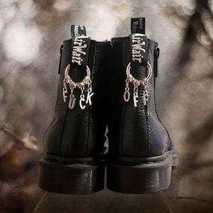 May include: Black leather ankle boots with AirWair tags and dangling charms spelling out "F*ck Off". The boots have a textured finish and a thick sole, with visible stitching details. The background is blurred.
