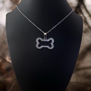 May include: A silver-toned necklace featuring a pendant shaped like a dog bone. The pendant is outlined with small, clear, sparkling stones set into the metal frame. The necklace hangs from a delicate, thin silver-colored metal chain.