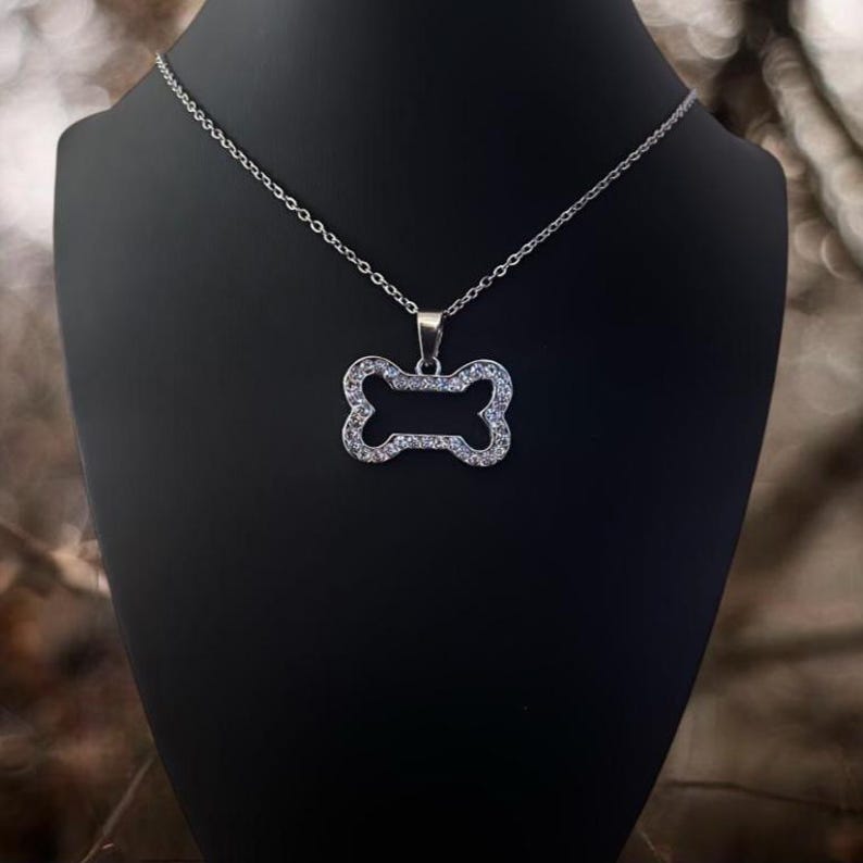 May include: A silver-toned necklace featuring a pendant shaped like a dog bone. The pendant is outlined with small, clear, sparkling stones set into the metal frame. The necklace hangs from a delicate, thin silver-colored metal chain.