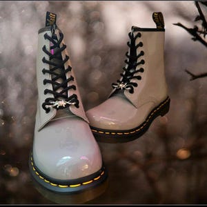 Może przedstawiać: Para szarych lakierowanych butów sznurowanych z czarnymi sznurówkami i srebrnymi okuciami. Buty mają żółte przeszycia wokół podeszwy.