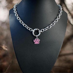Puede incluir: Collar de cadena plateada con un dije de huella de pata rosa brillante. El collar se exhibe en un maniquí negro. El dije de huella de pata es de color rosa claro.