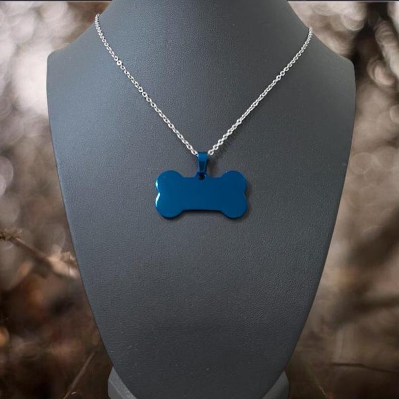 May include: A silver-toned metal chain necklace featuring a solid, smooth, dark blue pendant shaped like a dog bone. The pendant is suspended from a small metal bail attached to the center of the chain, creating a simple, clean, and uniform appearance.
