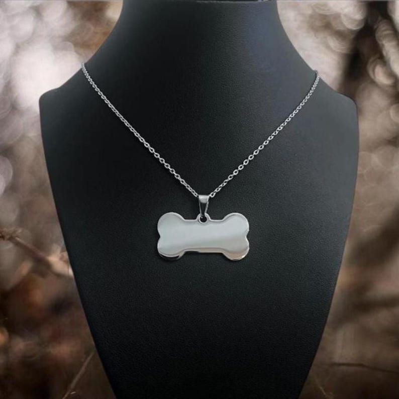 May include: A silver-toned metal necklace featuring a smooth, polished pendant shaped like a dog bone. The pendant is suspended from a delicate, thin link chain, creating a simple and clean design that rests against a dark display bust.