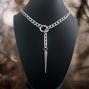 以下が含まれることがあります： 大きなリングと尖ったペンダントが付いた、シルバーカラーのチェーンネックレス。 黒いマネキンに飾られており、デザインと質感が際立っています。 チェーンのリンクは太く、ペンダントは円錐形です。