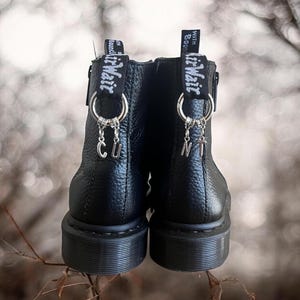 Puede incluir: Botas de tobillo de cuero negro con abalorios plateados que deletrean "CUNT" colgando de los cordones. Las botas tienen una cremallera negra y una correa negra con el texto "With God's Guidance" impreso en ella.