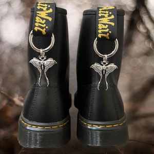 Könnte beinhalten: Ein Paar schwarze Stiefeletten mit gelben Nähten entlang der Sohle und einer schwarzen Fersenzuglasche mit gelbem AirWair-Text. Jeder Stiefel ist mit einem silberfarbenen Mottenanhänger aus Metall verziert, der an einem Ring hängt.