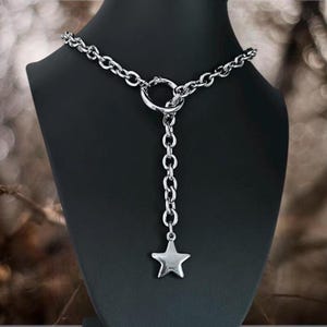 Könnte beinhalten: Eine silberfarbene Halskette mit Sternanhänger. Die Halskette hat ein Kettenmuster mit einem großen Ring in der Mitte und einer vertikalen Kette, die in einem fünfzackigen Stern endet. Der Schmuck wird auf einer schwarzen Schaufensterpuppe präsentiert.