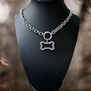 Puede incluir: Un collar plateado para perros con diseño de cadena. Presenta un colgante en forma de hueso incrustado con cristales transparentes. El collar se exhibe en un maniquí negro, mostrando su diseño y detalles.