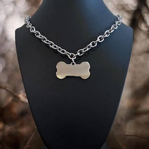 May include: A silver-colored dog collar with a bone-shaped tag. The collar is made of a chain link design. The bone-shaped tag is a light gold color and hangs from the center of the collar.