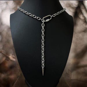 May include: A silver-toned metal chain necklace featuring a Y-shaped design with a central carabiner clasp connector. The necklace includes a vertical drop chain segment that terminates in a single, pointed metal spike pendant at the bottom end.
