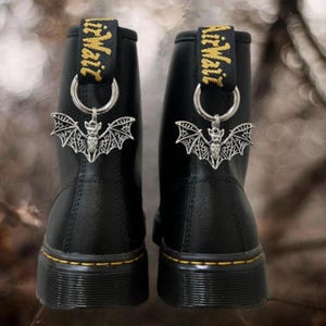 May include: A pair of black leather boots featuring yellow stitching along the sole and a black heel pull tab with the yellow text AirWair. Each boot is adorned with a silver-toned metal bat charm suspended from a circular ring on the heel tab.