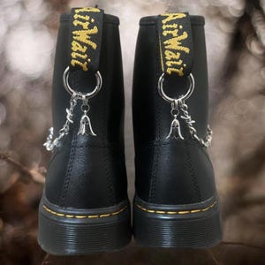 May include: Pair of black ankle boots with yellow stitching and soles. The boots have a pull tab with "Air Wair" in yellow lettering. Silver-colored chain and charm details hang from the back of the boots.